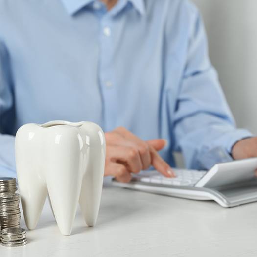 A person calculating costs while a ceramic tooth and coins sit in the foreground